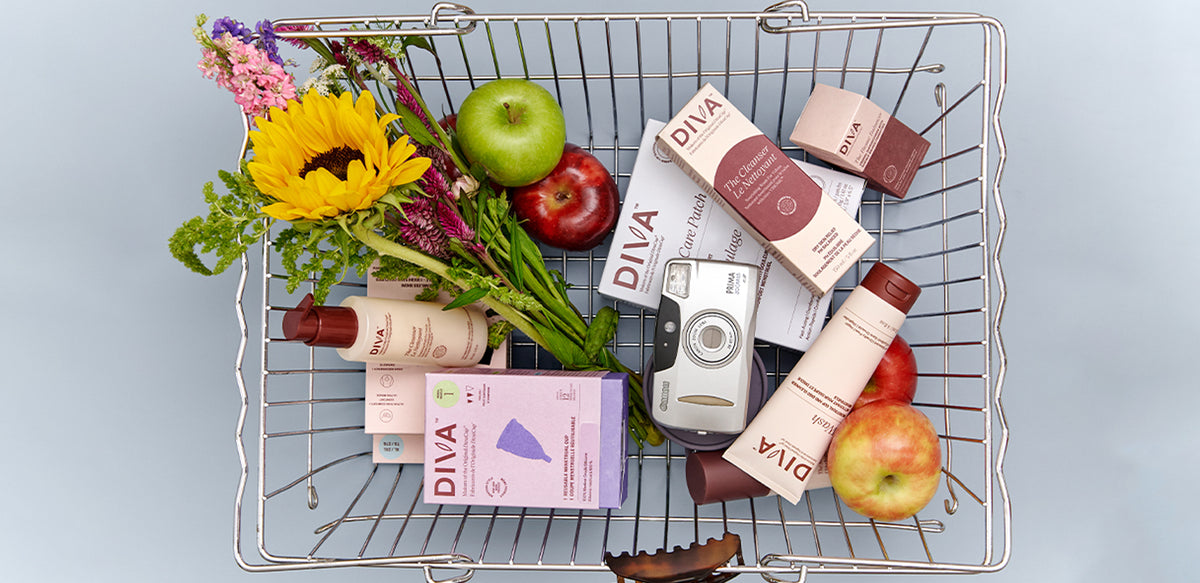 A top-down view of a metal wire shopping cart filled with assorted beauty products, flowers, and fruits against a light blue-gray background. Multiple DIVA-branded boxes and bottles are visible, along with apples, a green apple, and a camera.