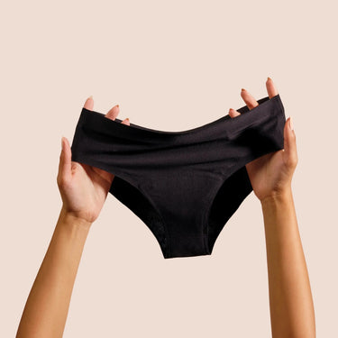Two hands hold up a pair of DIVA black reusable period underwear against a plain beige background. The underwear is stretched slightly to show its shape and seamless fabric.
