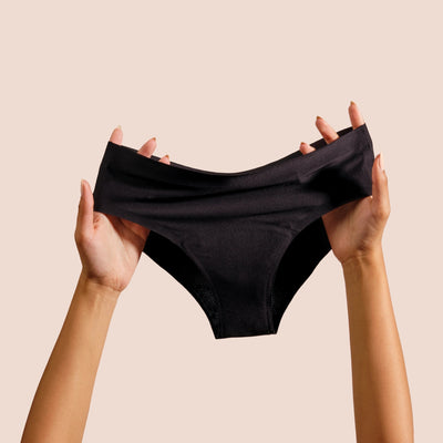 Two hands hold up a pair of DIVA black reusable period underwear against a plain beige background. The underwear is stretched slightly to show its shape and seamless fabric.